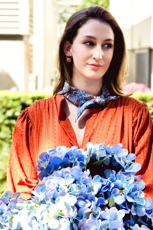 Woman wearing an orange blouse and blue neck scarf holding a bunch of blue flowers